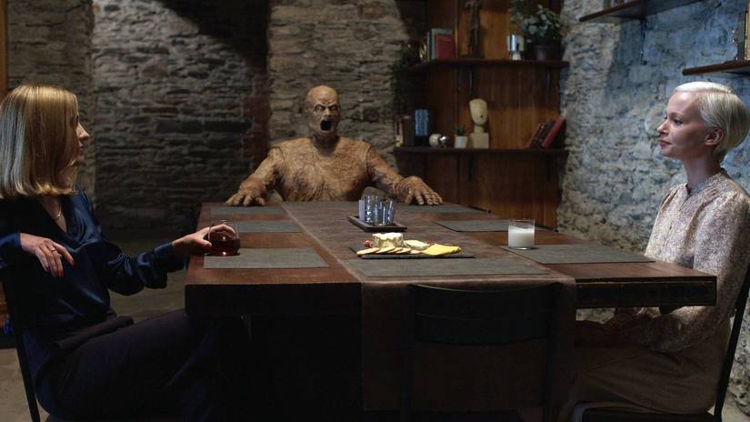 Two women sit across from each other at a table, looking calm, while a bald, naked, mottled-brown creature with its mouth open in a scream sits at the end of the table, fingers tented on the table's surface