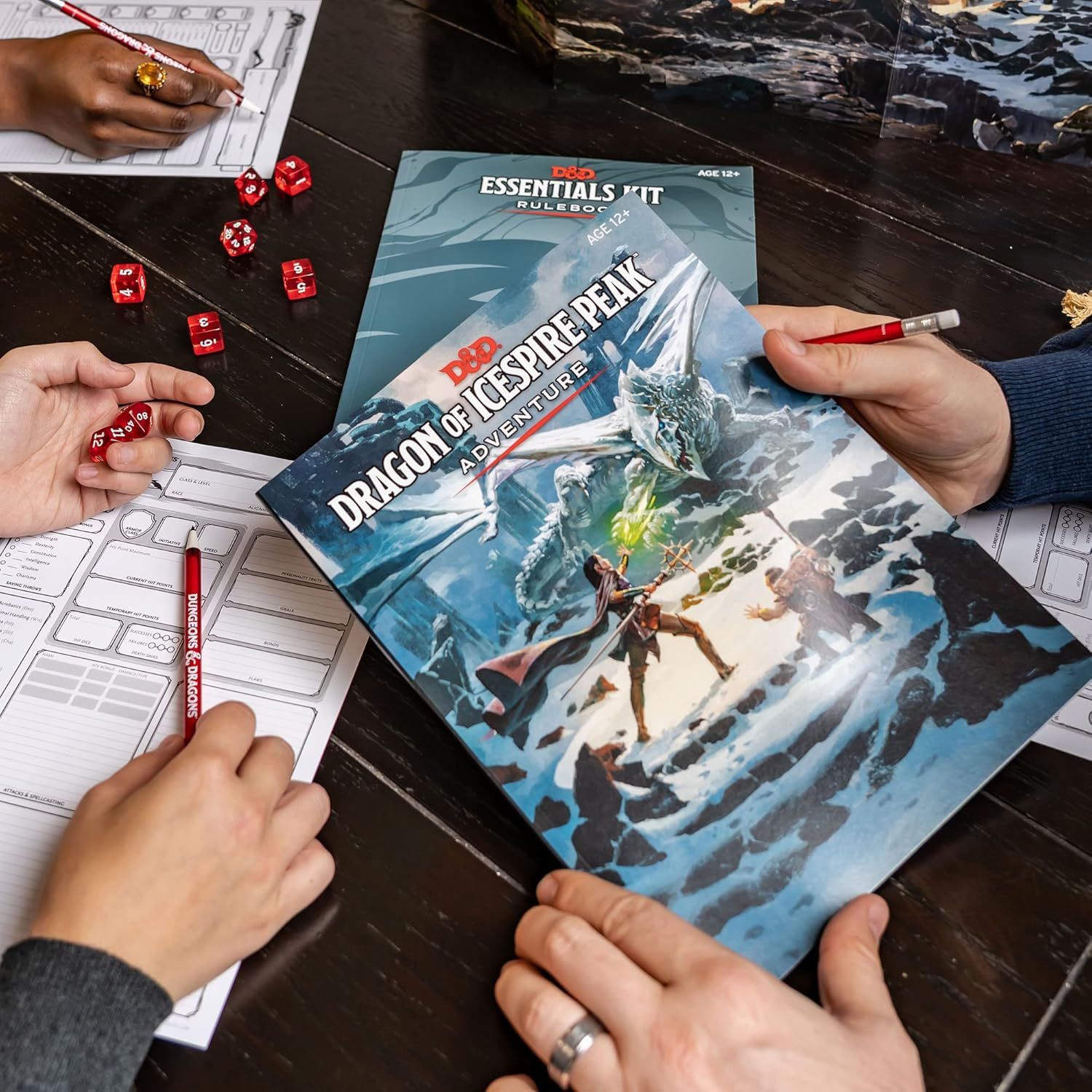 The contents of the D&D Essentials kit in action, with the adventure book held in hand in the foreground, the essentia rules just behind that, and several hands in various states of writing, rolling, and mulling over how to argue with the DM.