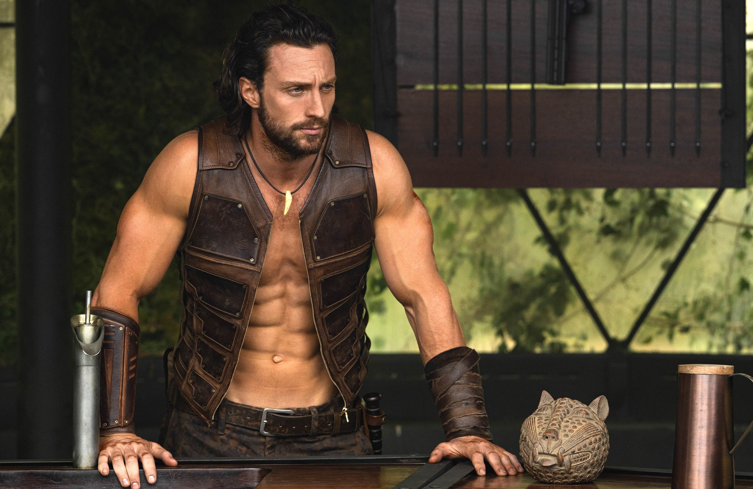 Kraven (Aaron Taylor-Johnson) stands with his hands on a railing, wearing a sleeveless, open leather vest and showing off more abs than seems humanly possible in Kraven the Hunter