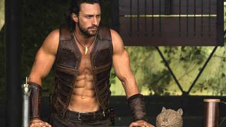 Kraven (Aaron Taylor-Johnson) stands with his hands on a railing, wearing a sleeveless, open leather vest and showing off more abs than seems humanly possible in Kraven the Hunter