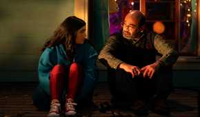 Kamala and her father sitting together on the floor. Kamala has her knees pulled to her chest and looks a bit nervous, while her father looks understanding. From Ms. Marvel.