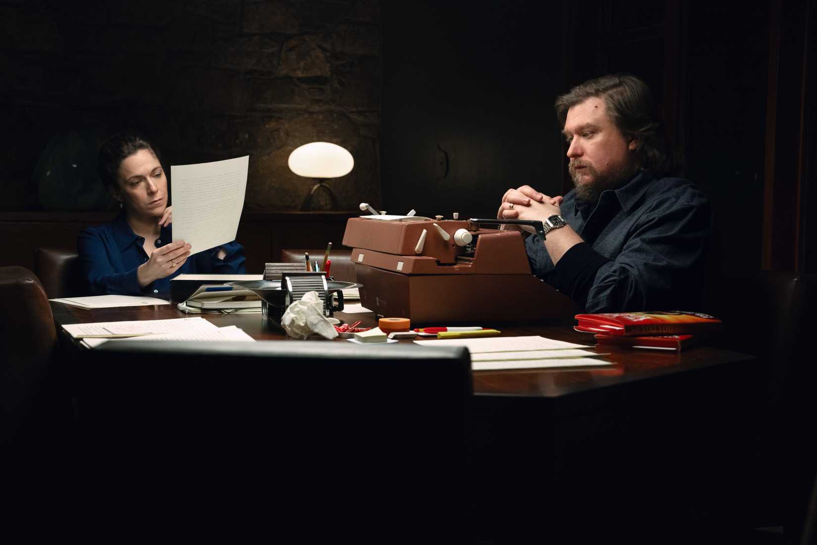 In a still from Severance, Devon (Jen Tullock) reads a paper while Ricken (Michael Chernus) sits in front of a typewriter with his hands folded