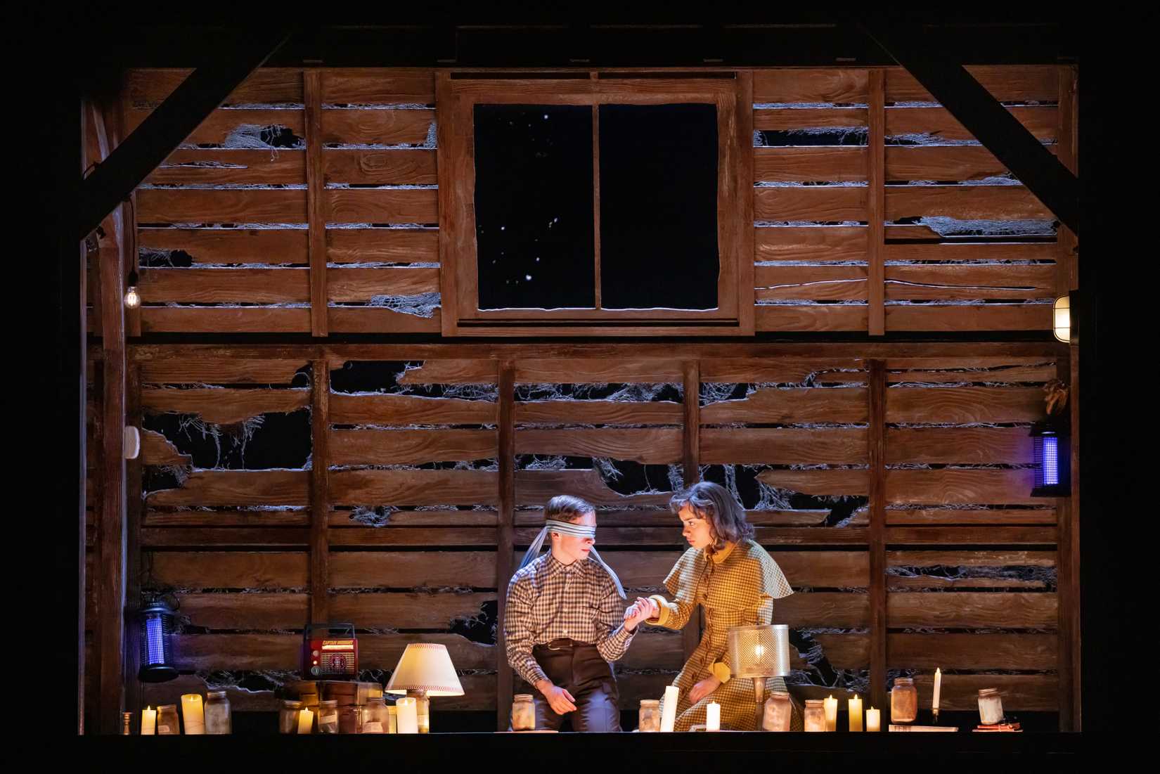 Henry, blindfolded, holding hands with Patty Newby in an attic full of candles. From Stranger Things: The First Shadow