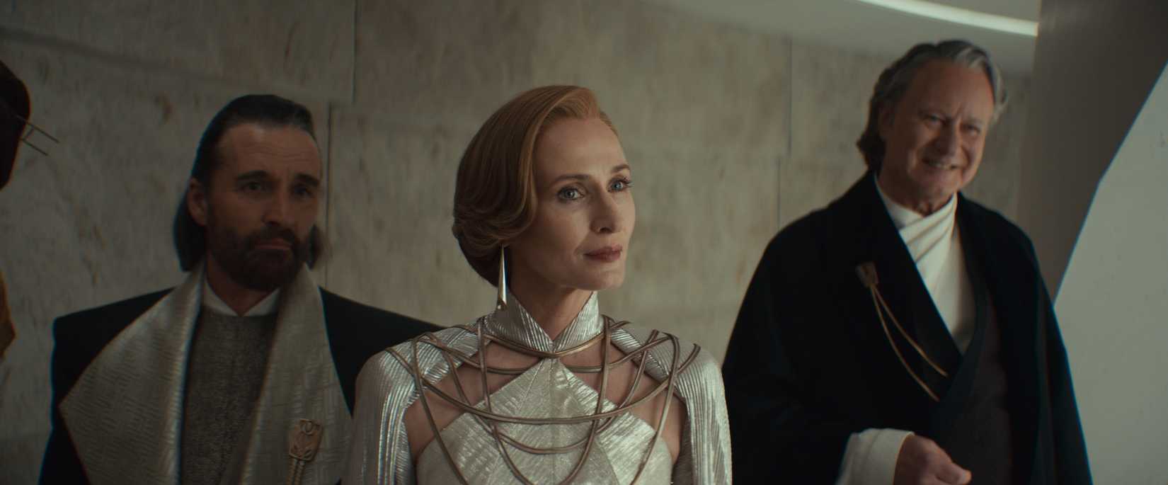 A shot of Andor season 2. It features Perrin Fertha, a smug-looking human male with a goatee and pushed back brown hair. He wears fancy garb. Next to him is Mon Mothma, who wears a white, crystal-like dress with fancy earrings. She sports short, pushed back ginger hair and looks serious. To her right is Luthen Rael, wearing a black and white fancy robe. He looks pleased as punch.