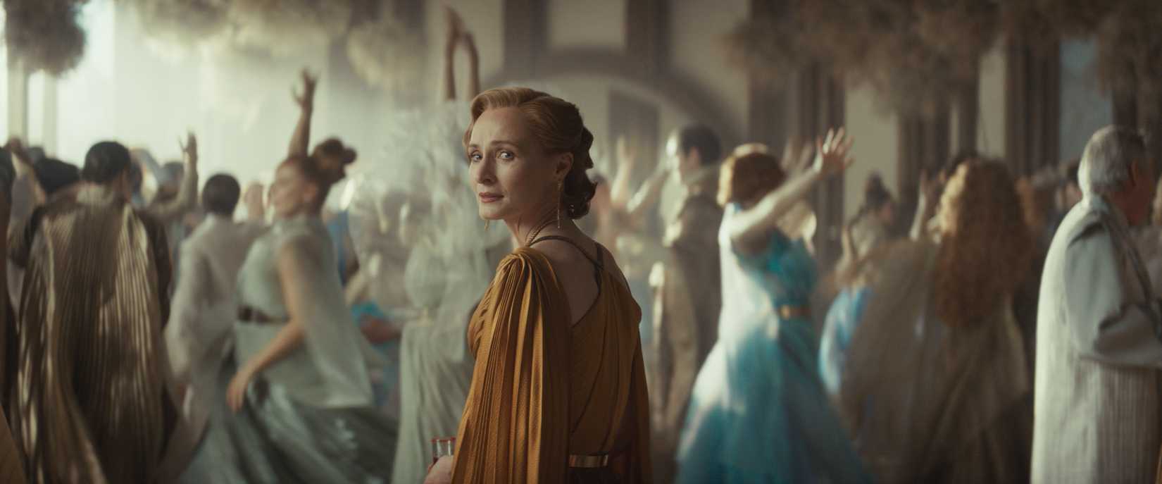Mon Mothma (Genevieve O'Reilly) in Andor season 2. She looks over her shoulder sadly, standing in a ballroom with wildly swirling wedding dancers behind her.