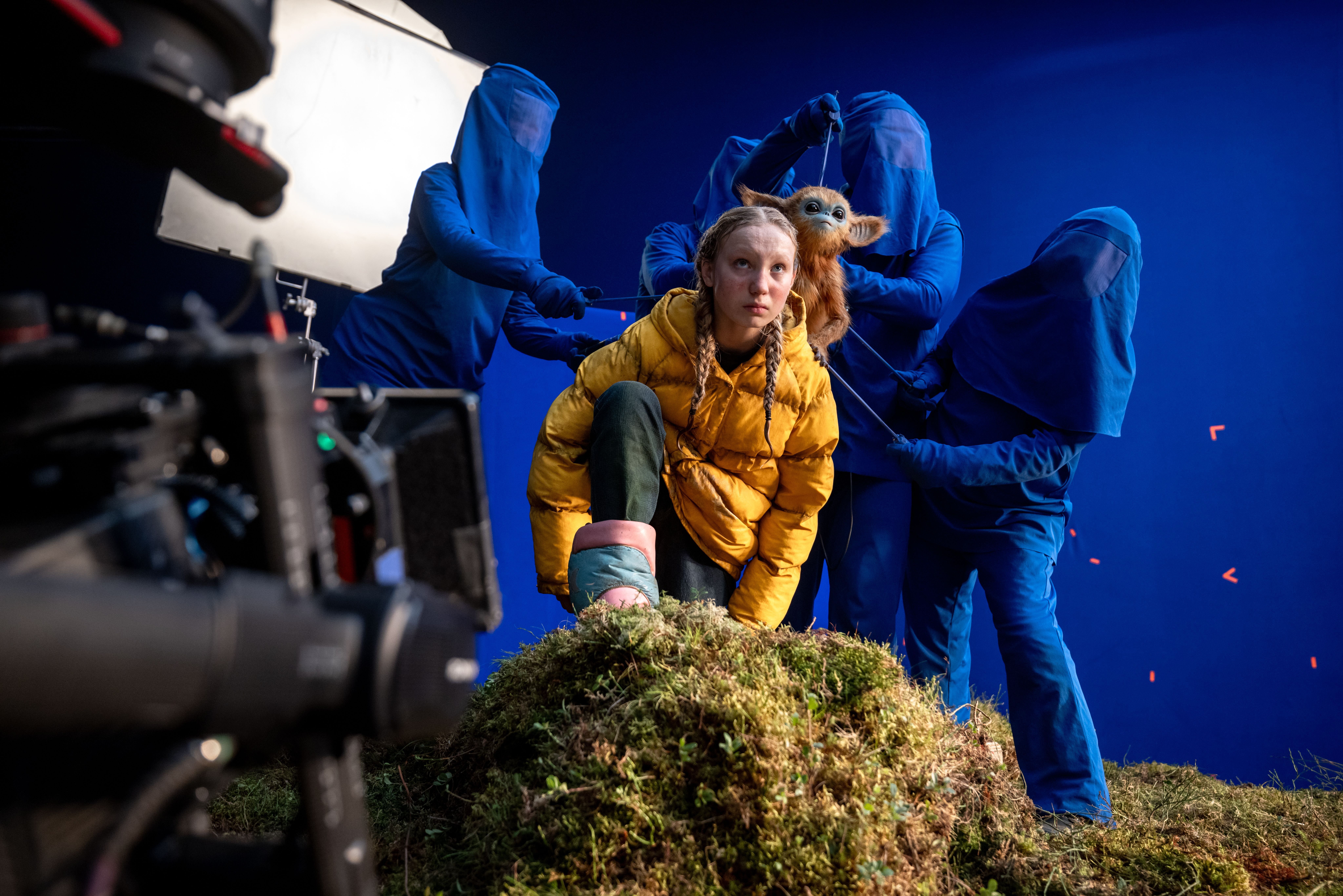 A girl in a yellow coat crouches in front of a blue screen. There is a tiny monkey-like puppet on her shoulder, operated by a team of people behind her, draped and veiled in dark blue cloth. From The Legend of Ochi