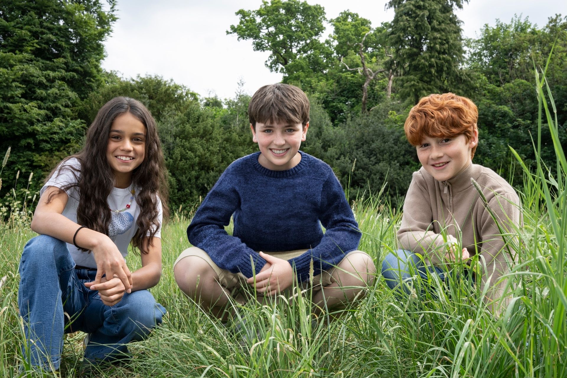 A photo of actors Arabella Stanton, Dominic McLaughlin, and Alastair Stout squatting in a field of grass, for HBO’s new Harry Potter TV series
