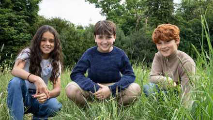 A photo of actors Arabella Stanton, Dominic McLaughlin, and Alastair Stout squatting in a field of grass, for HBO’s new Harry Potter TV series