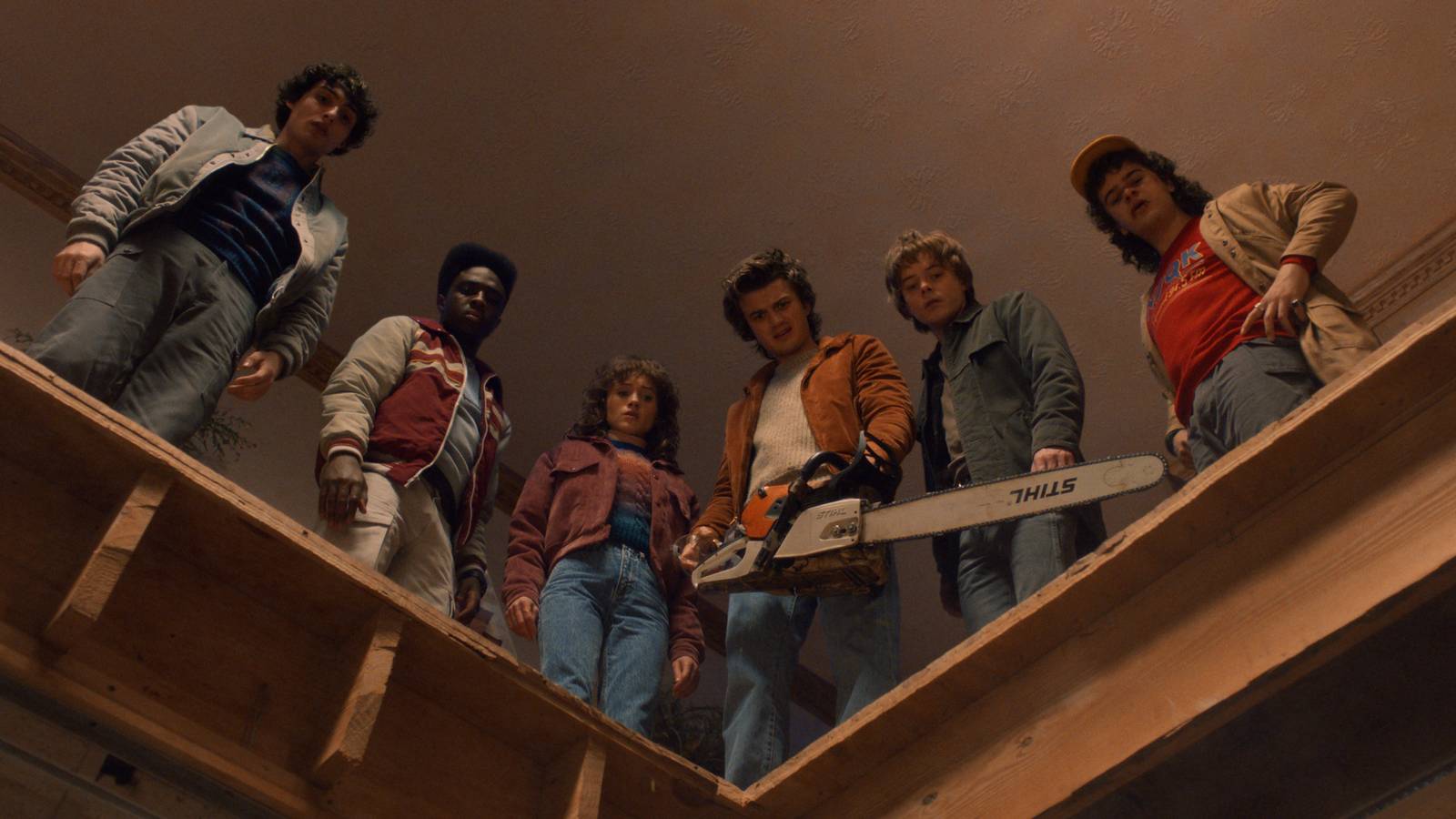 Finn Wolfhard as Mike Wheeler, Caleb McLaughlin as Lucas Sinclair, Natalia Dyer as Nancy Wheeler, Joe Keery as Steve Harrington, Charlie Heaton as Jonathan Byers, and Gaten Matarazzo as Dustin Henderson stand in front of a hole in a building while Steve holds a chainsaw in Stranger Things