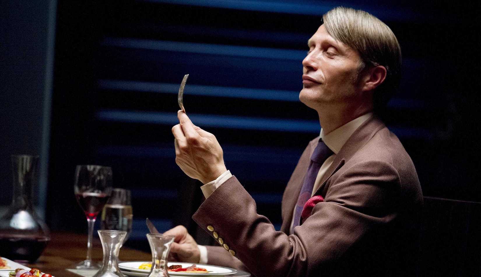 Hannibal Lecter (Mads Mikkelsen) sits at a place setting, examining a fork, in season 2 of Hannibal