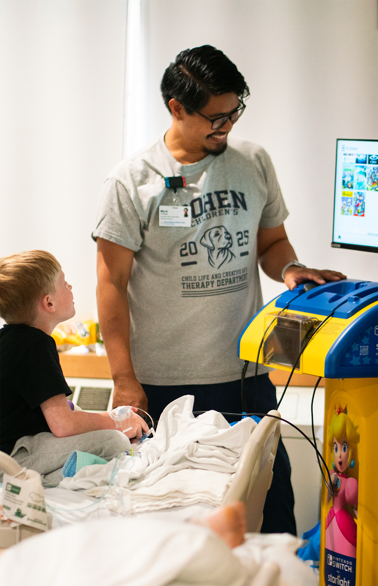 A small blond boy is in a hospital bed playing a video game, watched over by a smiling dark-haired aide.