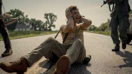 A skinny teenager sits in the road, clutching his head and crying, as soldiers carrying rifles approach him in The Long Walk