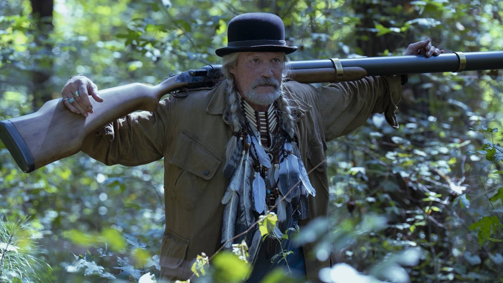 A man in a duster and bowl hat holding a rifle behind his back in a wooded setting from Peacemaker season 2, episode 5