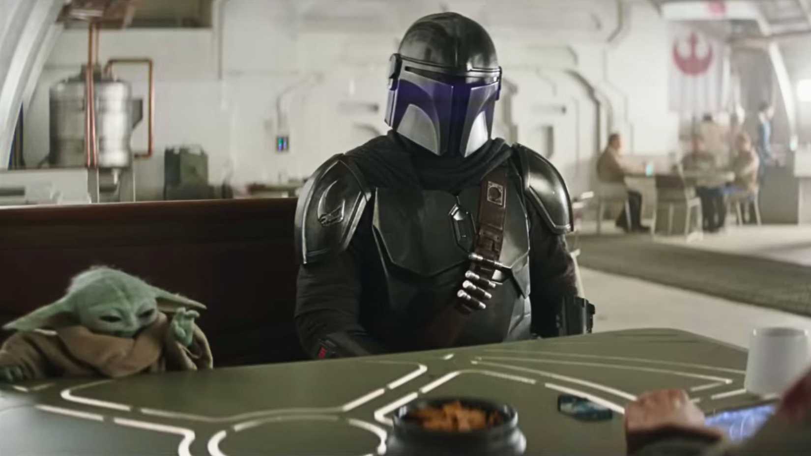 An image from The Mandalorian and Grogu. Both Grogu and Din Djarin sit at a table, with Grogu's hand outstretched.
