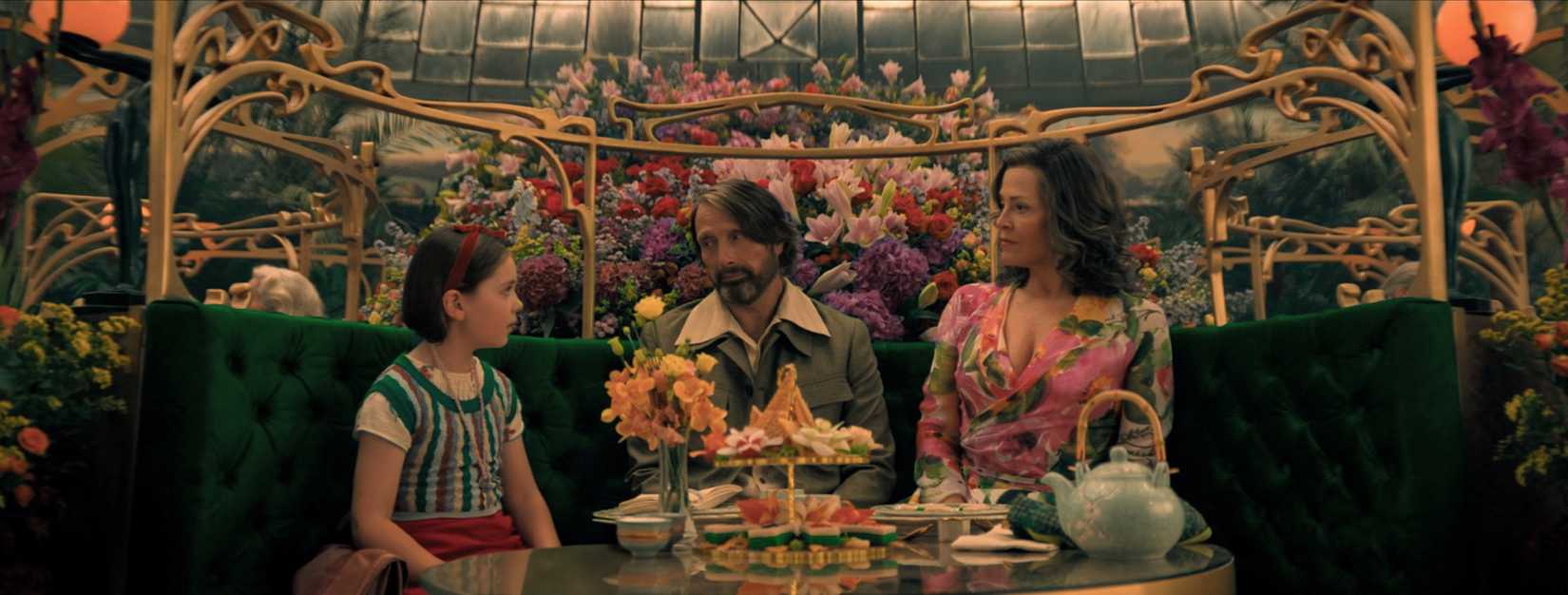 Image from Dust Bunny featuring Sigourney Weaver, Mads Mikkelsen and Sophie Sloan. The three of them sit in a cafe full to the brim with flowers. A tea set sits in front of the three of them.