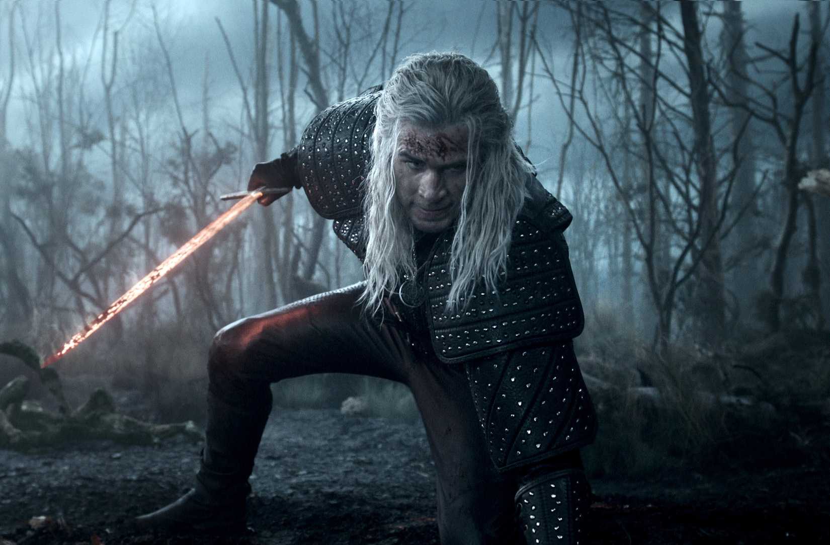 Liam Hemsworth as The Witcher crouching with a flaming sword in a blue-hued woods