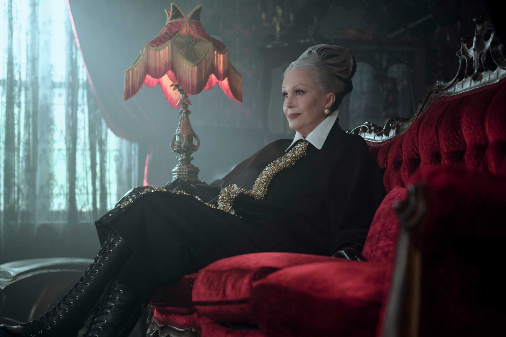 Wednesday image featuring Hester Addams. She sits on a lush, red sofa.