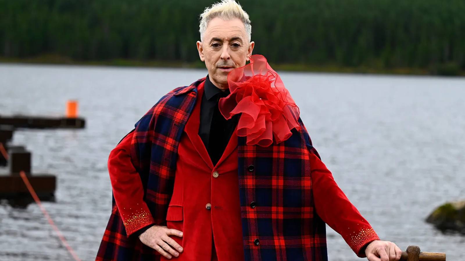 Alan Cumming as host of The Traitors, wearing a red outfit with a plaid scarf and dramatic ruffle