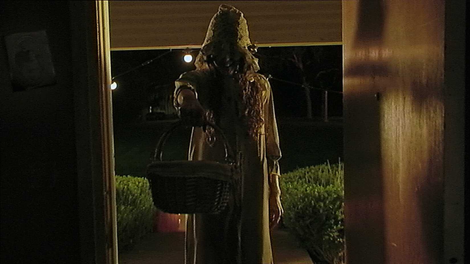 A spooky witch with a creepy grin blocks an open doorway out of a house in the V/H/S Halloween short "Coochie Coochie Coo"