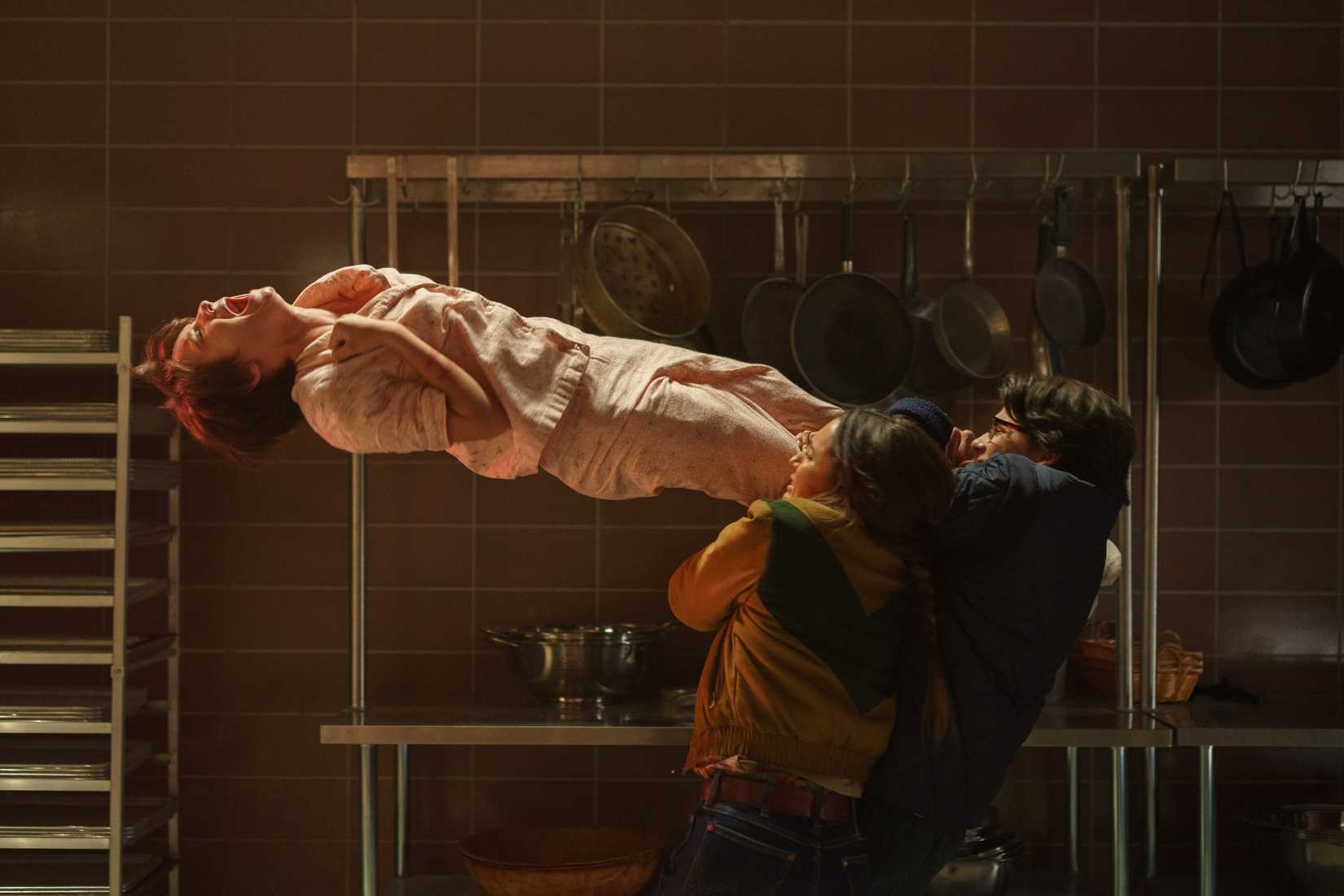 Gwen (Madeleine McGraw), in pink pajamas, floats on her back in the air, screaming and clutching at her throat, as several people cling to her legs and try to pull her down in Black Phone 2