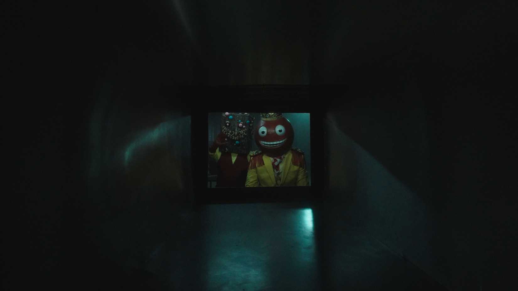 Two surreal candy-headed creatures peer from a small window at the end of a metal tunnel