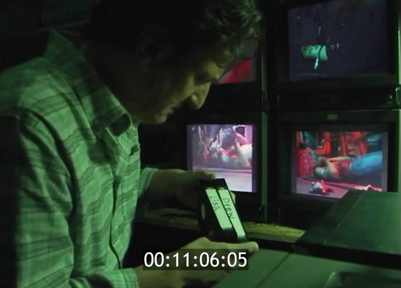 A man looks at a pair of videotapes marked "Drew" and "Lisa" as bloody scenes play out on the cameras behind him in V/H/S Halloween's "Kidprint" segment