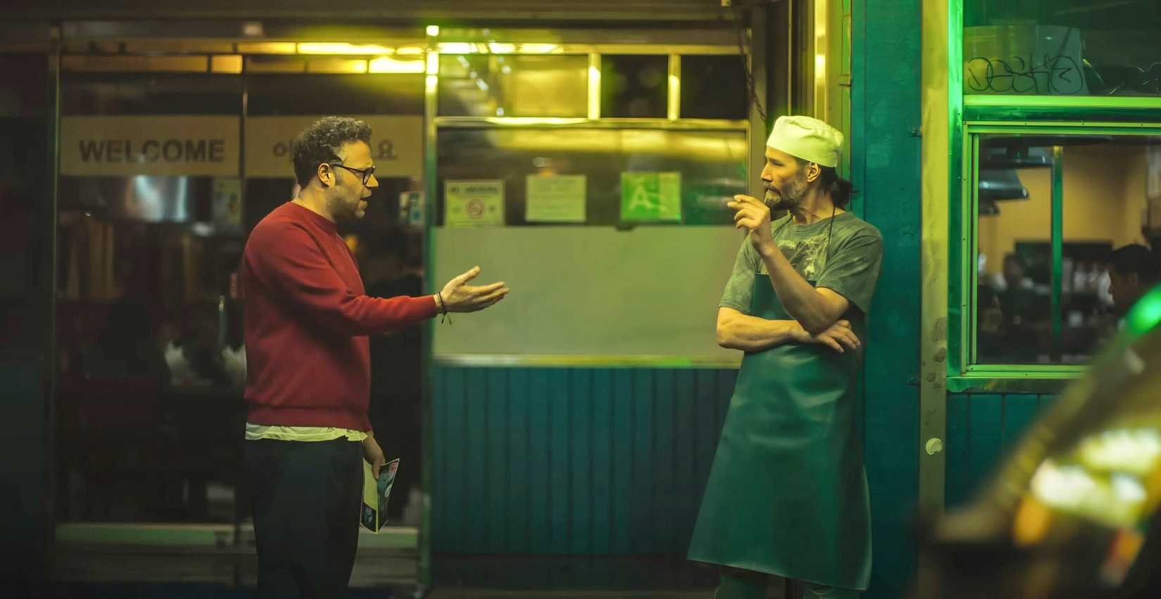 Jeff (Seth Rogen) confronts Gabriel (Keanu Reeves) in the unflattering green outdoor light of a strip-mall Korean restaurant. Reeves has his hair pulled back in a ponytail, is wearing an apron and paper food-service cap, and is smoking.