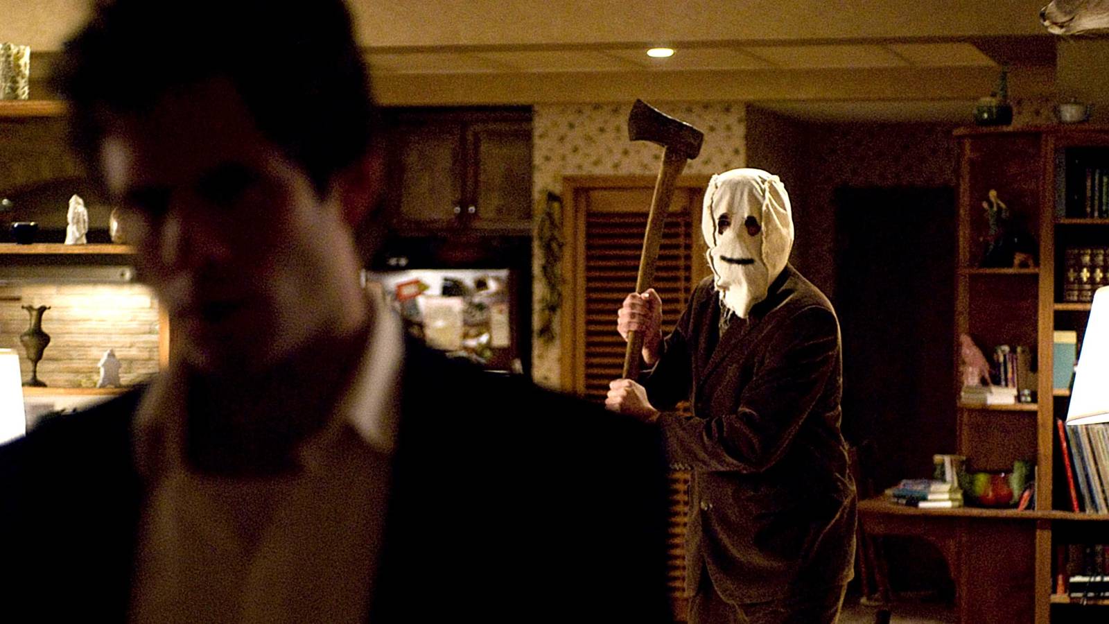 Masked figure with holes for eyes and a drawn smile raising an axe behind an unsuspecting man — still from The Strangers (2008)