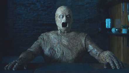 A horrifying golem posed to be sitting at a table with its mouth open in Oddity