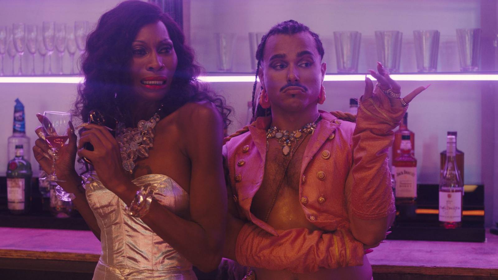 Dominique Jackson in a silver bustier and Tomás Matos shirtless in an open salmon leather vest glance off-screen at a bar in Queens of the Dead