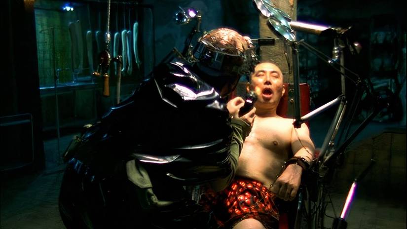 A man strapped to a chair; another with a helmet shines a light in his face, Save the Green Planet!