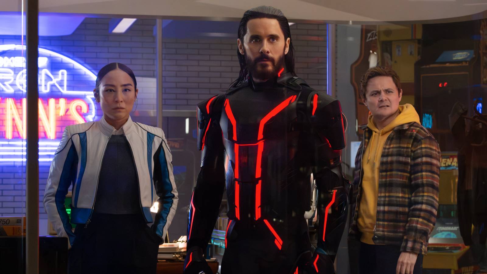 Eve Kim (Greta Lee), Ares (Jared Leto) and Seth Flores (Arturo Castro) stand together facing the camera in Flynn's Arcade in Tron: Ares