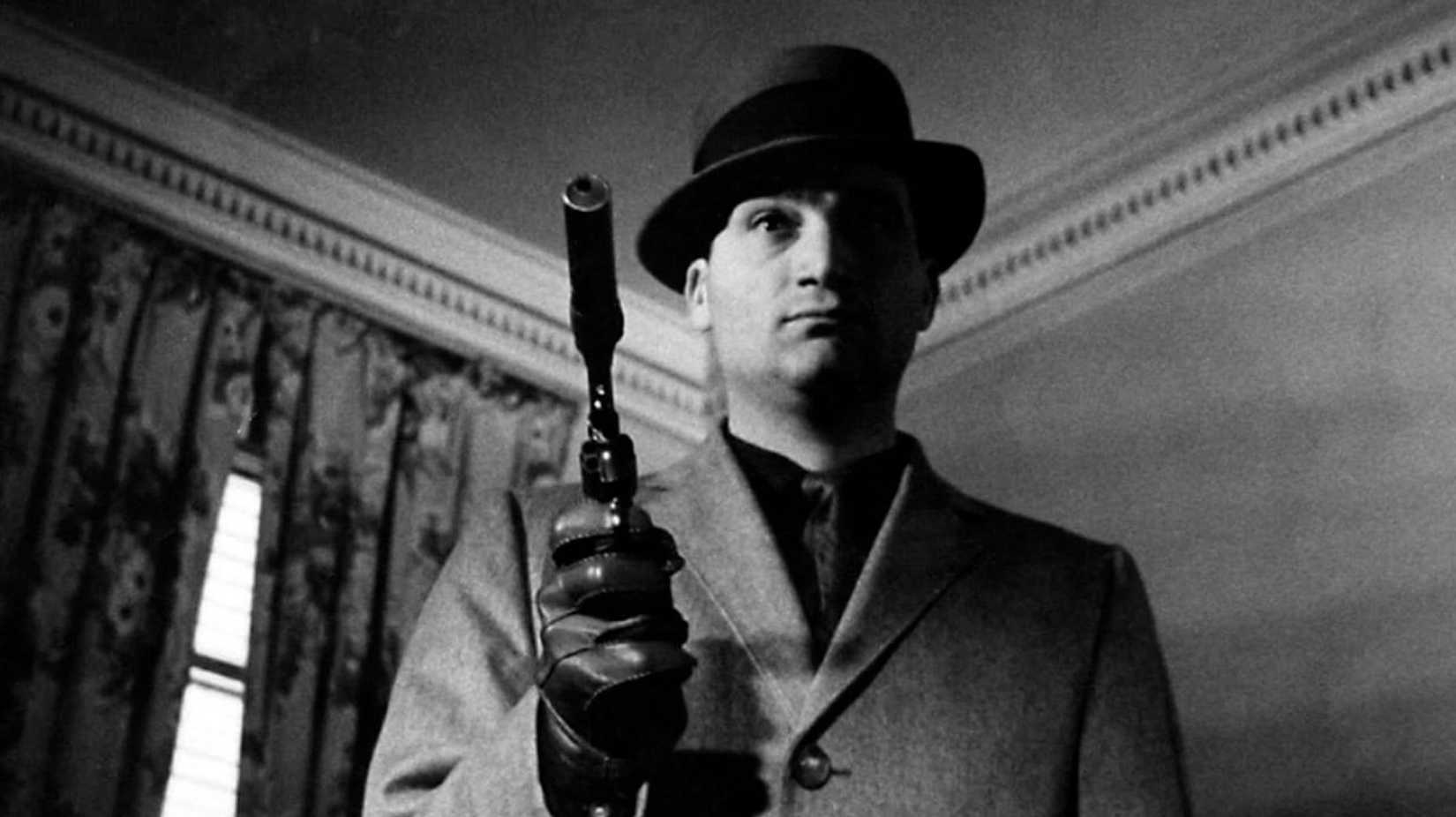 A man in a fedora and overcoat holds up a pistol equipped with a silencer in Blast of Silence