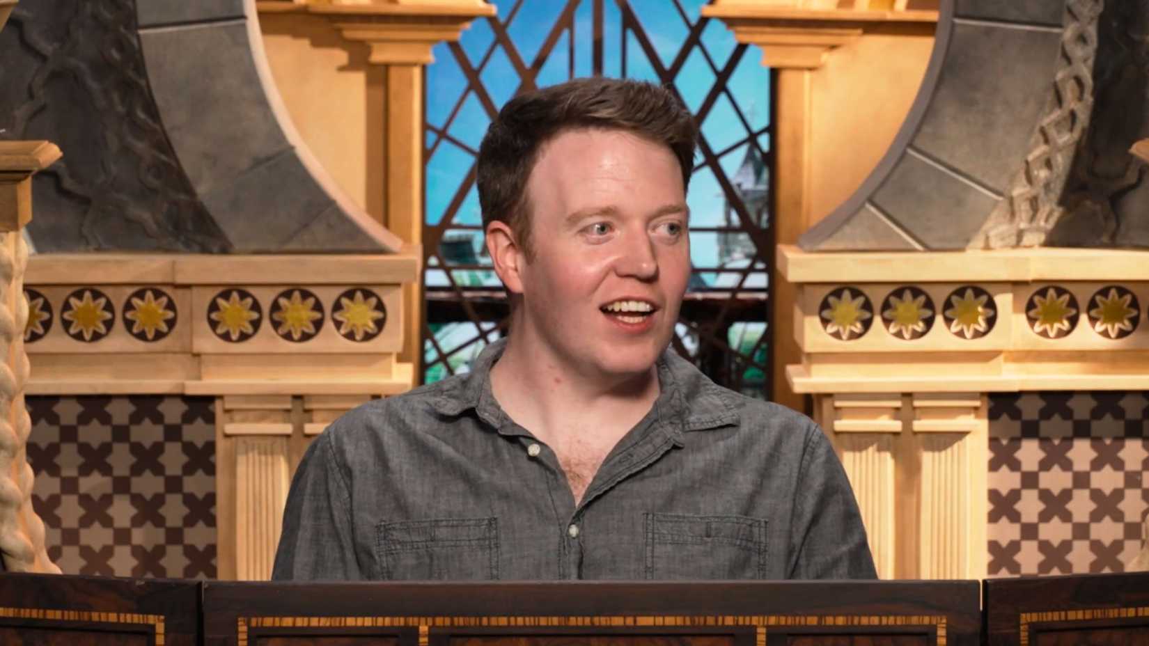 An image of Brennan Lee Mulligan during Critical Role's Cooldown for episode 5. Brennan wears a grey, open-buttoned shirt and is looking off to the side with a smile.