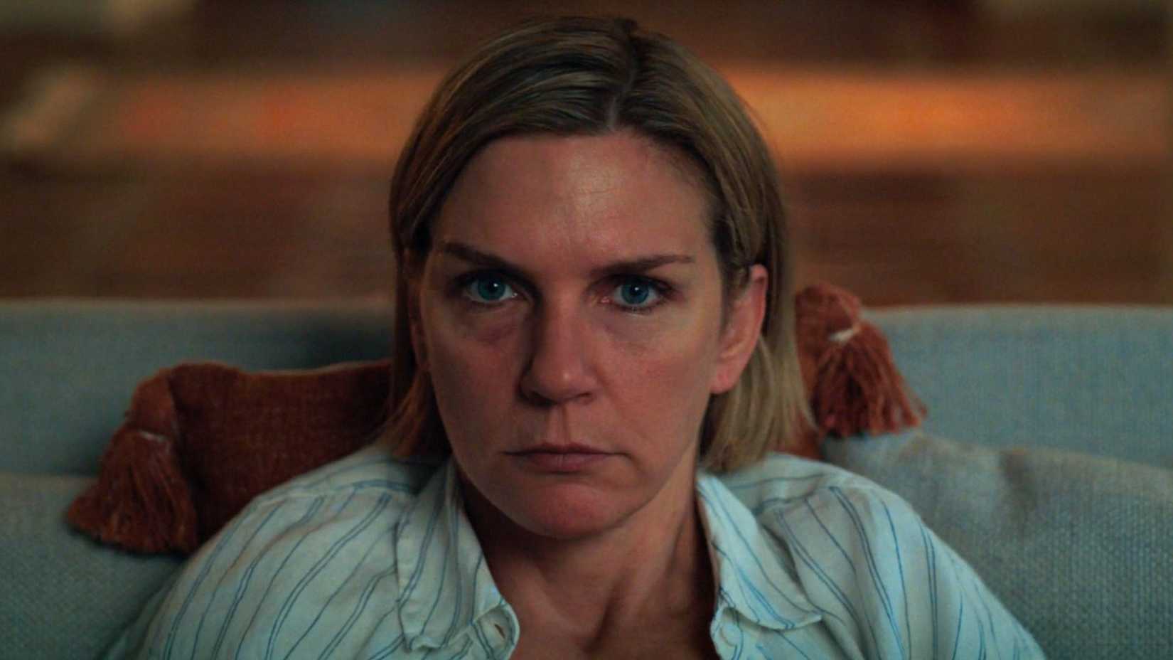 Carol (Rhea Seehorn) olha diretamente para frente enquanto a TV passa. Ela está no sofá, com uma almofada atrás dela.