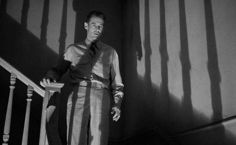 A man stands on the landing of a set of stairs, in shadow, with vertical bars of darkness across his body, in 1947's Crossfire
