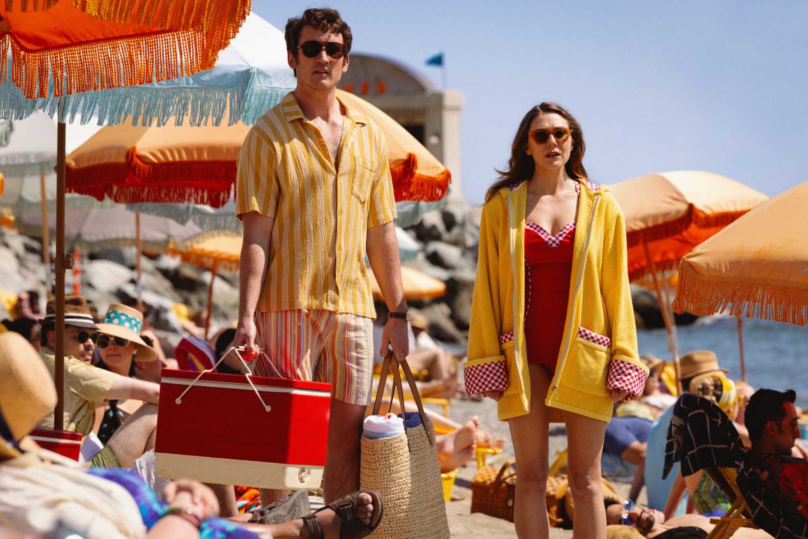 Larry and Joan (Miles Teller, Elizabeth Olsen) stand on a beach in a sunny afterlife, looking bemused, in 2025's Eternity