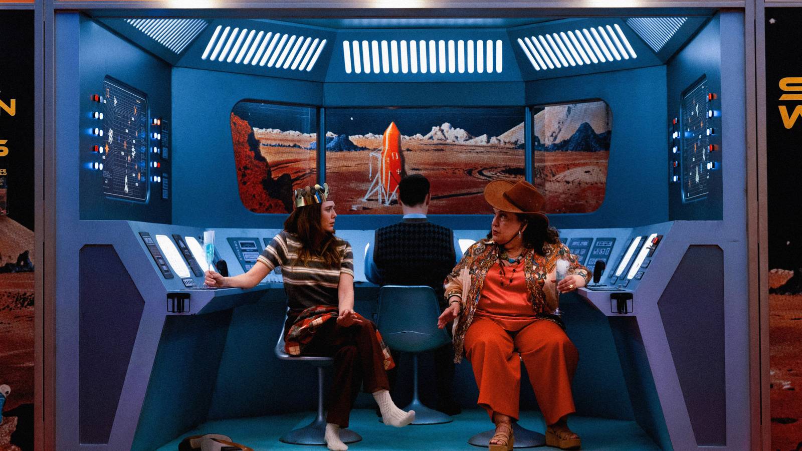 Two women (Elizabeth Olsen, Olga Merediz) sit in a cheap-looking mockup of a retro spaceship control booth, holding bright blue cocktails, in Eternity