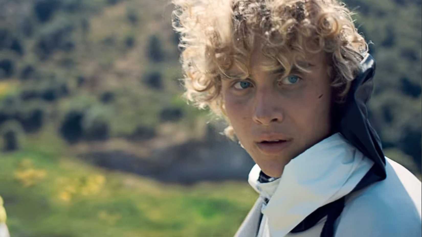 A young man with blonde curly hair looks over, surrounded by lush green fields.
