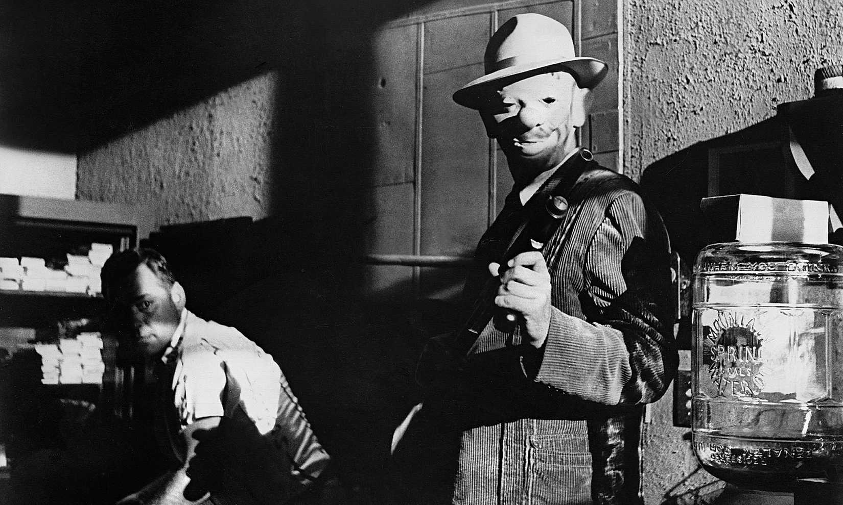 A man in a clown mask brandishes a gun while another man behind him sinks into the shadows in Stanley Kubrick's The Killing