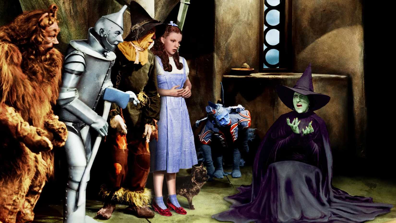 A Bruxa Má do Oeste grita e derrete enquanto Dorothy, o Leão Covarde, o Homem de Ferro e o Espantalho assistem ao Mágico de Oz de 1939.