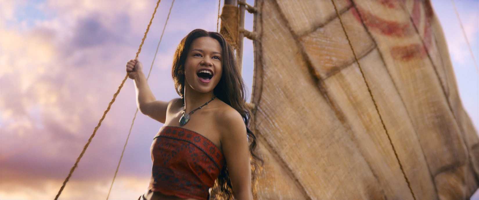 Catherine Laga'aia as Moana. She holds onto the rope of a sail and leans forward, mouth open in song.