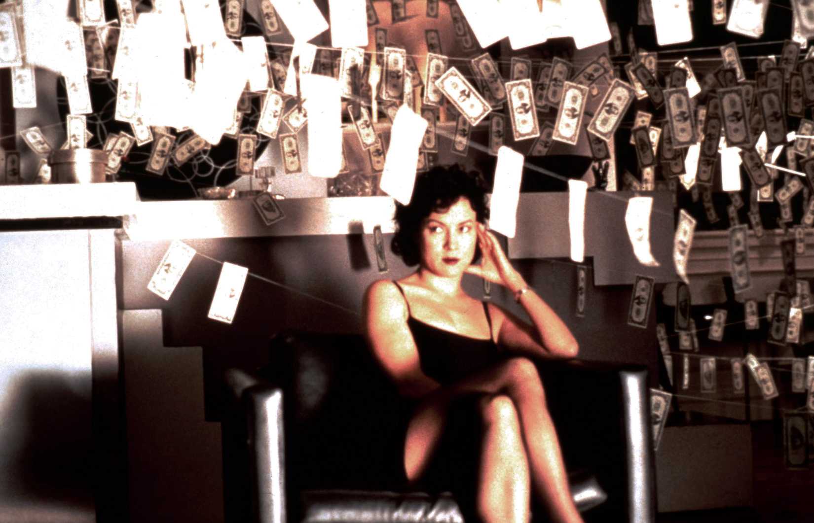 Jennifer Tilly, wearing a little black dress, sits cross-legged in a room strung with money hung from lines like laundry in Bound
