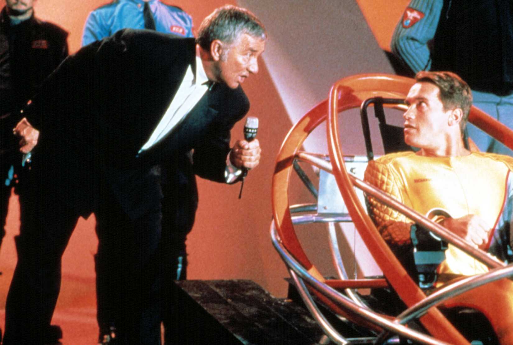 Game-show host Killian (Richard Dawson) leans down with a microphone to speak to Ben Richards (Arnold Schwarzenegger, wearing a golden futuristic bodysuit and strapped into a high-tech sled) in 1987's The Running Man