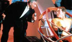 Game-show host Killian (Richard Dawson) leans down with a microphone to speak to Ben Richards (Arnold Schwarzenegger, wearing a golden futuristic bodysuit and strapped into a high-tech sled) in 1987's The Running Man