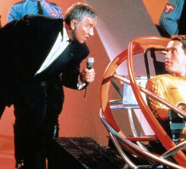Game-show host Killian (Richard Dawson) leans down with a microphone to speak to Ben Richards (Arnold Schwarzenegger, wearing a golden futuristic bodysuit and strapped into a high-tech sled) in 1987's The Running Man