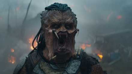 A Predator roars with his while standing in front of fiery wreckage in Predator: Badlands