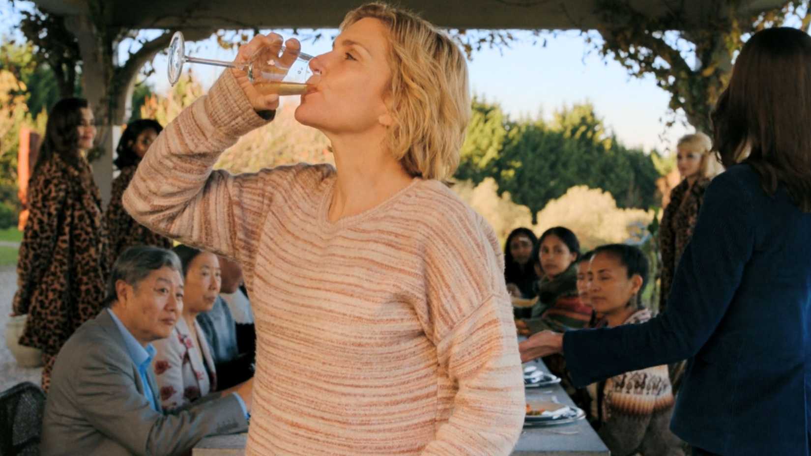 A screenshot from Pluribus episode 2 featuring Rhea Seehorn as Carol Sturka. She drinks from a wine glass, surrounded by her fellow immunes and a gaggle of hiveminds.
