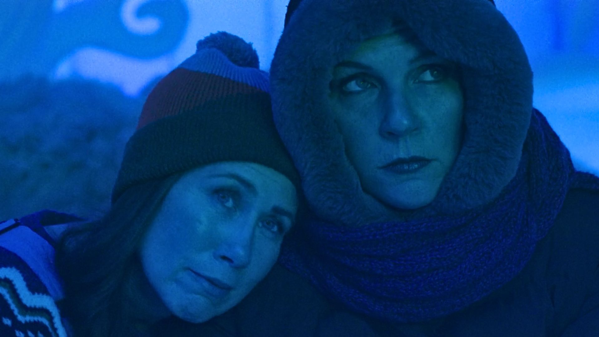 A screenshot from Pluribus featuring Miriam Shor as Helen and Rhea Seehorn as Carol Sturka. The two sit together, bundled up in coats, Helen's head on Carol's shoulder. Helen looks content. Carol looks grumpy. This is not new.