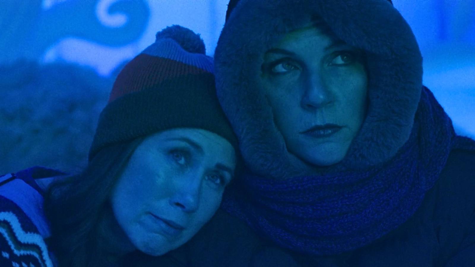 A screenshot from Pluribus featuring Miriam Shor as Helen and Rhea Seehorn as Carol Sturka. The two sit together, bundled up in coats, Helen's head on Carol's shoulder. Helen looks content. Carol looks grumpy.